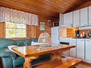 A cozy cabin interior with a wooden ceiling, a green sofa, a wooden dining table, and a kitchen area with white cabinets and appliances.