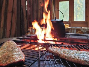 A grill with two pieces of meat cooking over an open flame, with a pot on a separate burner in the background.