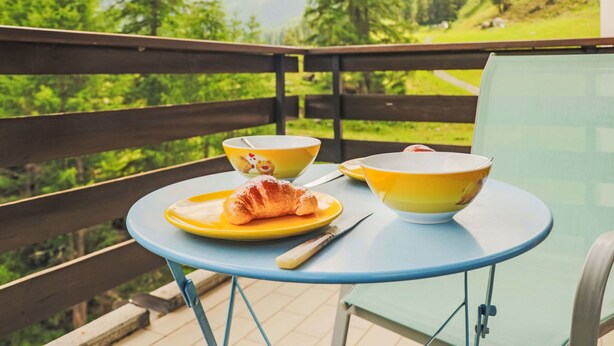 Een balkon met een tafel gedekt voor het ontbijt, inclusief croissants en kommen, met uitzicht op een bergachtig landschap.