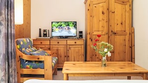 Een kamer met een houten kast, een televisie waarop een fietsevenement te zien is, een bank met een fruitmotief en een tafel met een bloevas.