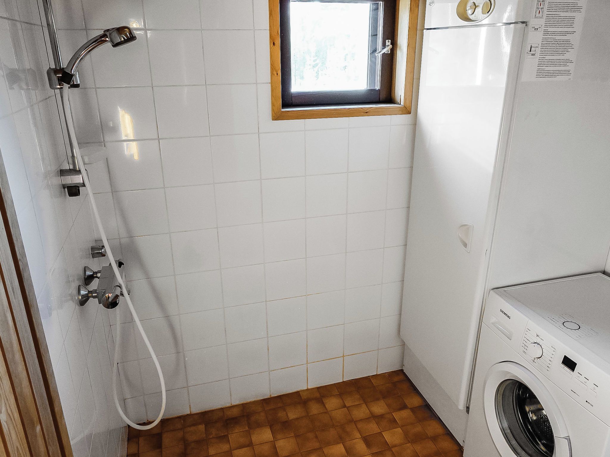 Une petite salle de bain propre, avec des carreaux blancs, une fenêtre en bois, une douche et un lave-linge.