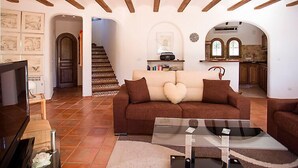 Un ampio soggiorno con un divano marrone, un tavolino da caffè in vetro e una televisione. Si nota una scala che conduce a un'altra stanza e una zona cucina con un arco.