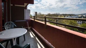 Een balkon met een tafel en stoelen, uitzicht op gebouwen en bomen, en een heldere lucht.