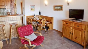 Un salón acogedor con un mueble bajo de madera para el televisor, una mesa de comedor con sillas, y un sillón rojo a cuadros.