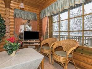 A cozy cabin interior with a wooden interior, a television on a desk, wicker chairs, and a view of a snowy landscape through large windows.
