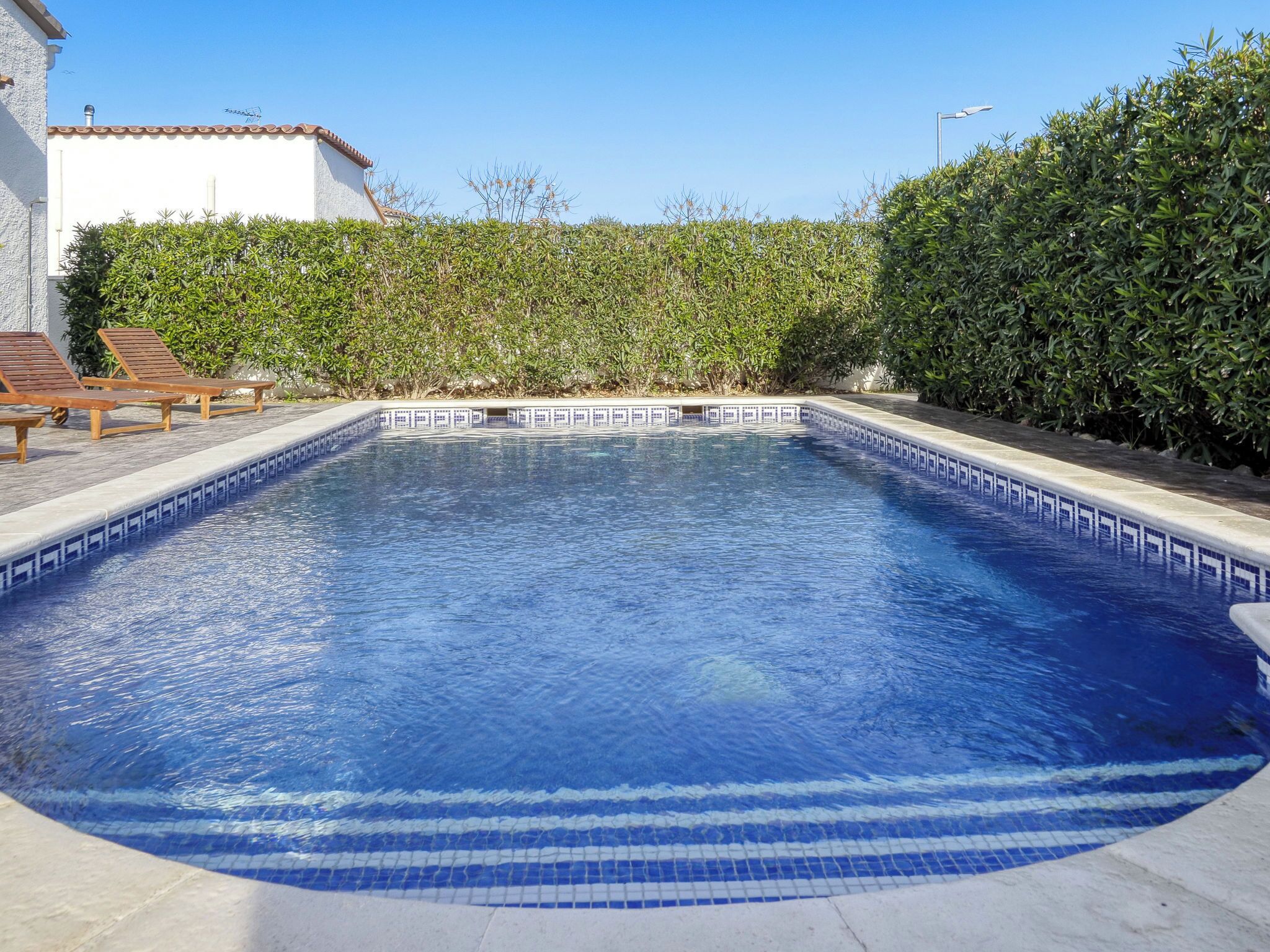 Une piscine dont le bord est recouvert de carreaux, entourée d’une haie et bordée d’un bâtiment à la façade blanche et au toit recouvert de tuiles.
