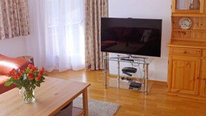 A living room with a flat-screen TV on a glass stand, a wooden cabinet with glass doors, a wooden table with a vase of flowers, and a red sofa. - Chesa Sur Val 22 by Interhome (St. Moritz)