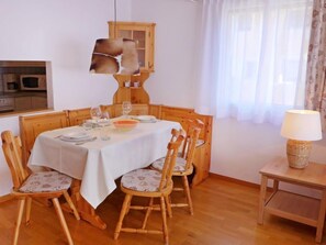 A dining area with a wooden table set for four, surrounded by four wooden chairs with cushioned seats. A wooden cabinet with glass doors is above the table, and a lamp is on a side table to the right. - Chesa Sur Val 22 by Interhome (St. Moritz)