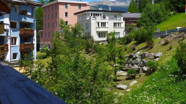 A hillside residential area with multi-story buildings, including a pink and white building with balconies, surrounded by greenery and a mountain backdrop. - Chesa Sur Val 22 by Interhome (St. Moritz)