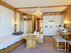 Une pièce confortable avec un lit, une table en bois et un banc. On y trouve également une petite cuisine équipée d’une cuisinière et d’un évier.