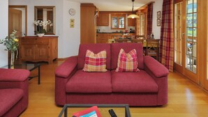 Una sala de estar con un sofá rojo, una mesa de centro con un libro y un control remoto, armarios de madera, una cocina al fondo y un amplio ventanal con cortinas.