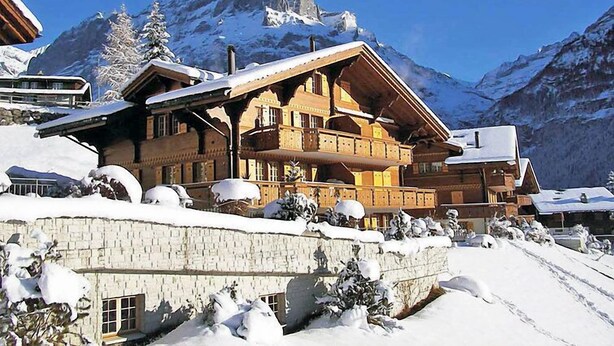 Un edificio de estilo chalé con techo pronunciado, balcones de madera y base de piedra, rodeado de montañas nevadas.