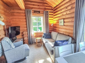 Et interiør af en træhytte med en sofa, to lænestole, et lille bord, et fjernsyn og et vindue med gardiner.