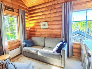 L’intérieur d’un chalet chaleureux, avec un mur en rondins de bois, un canapé avec des coussins, une fenêtre avec des rideaux et une vue sur un pont à l’extérieur.