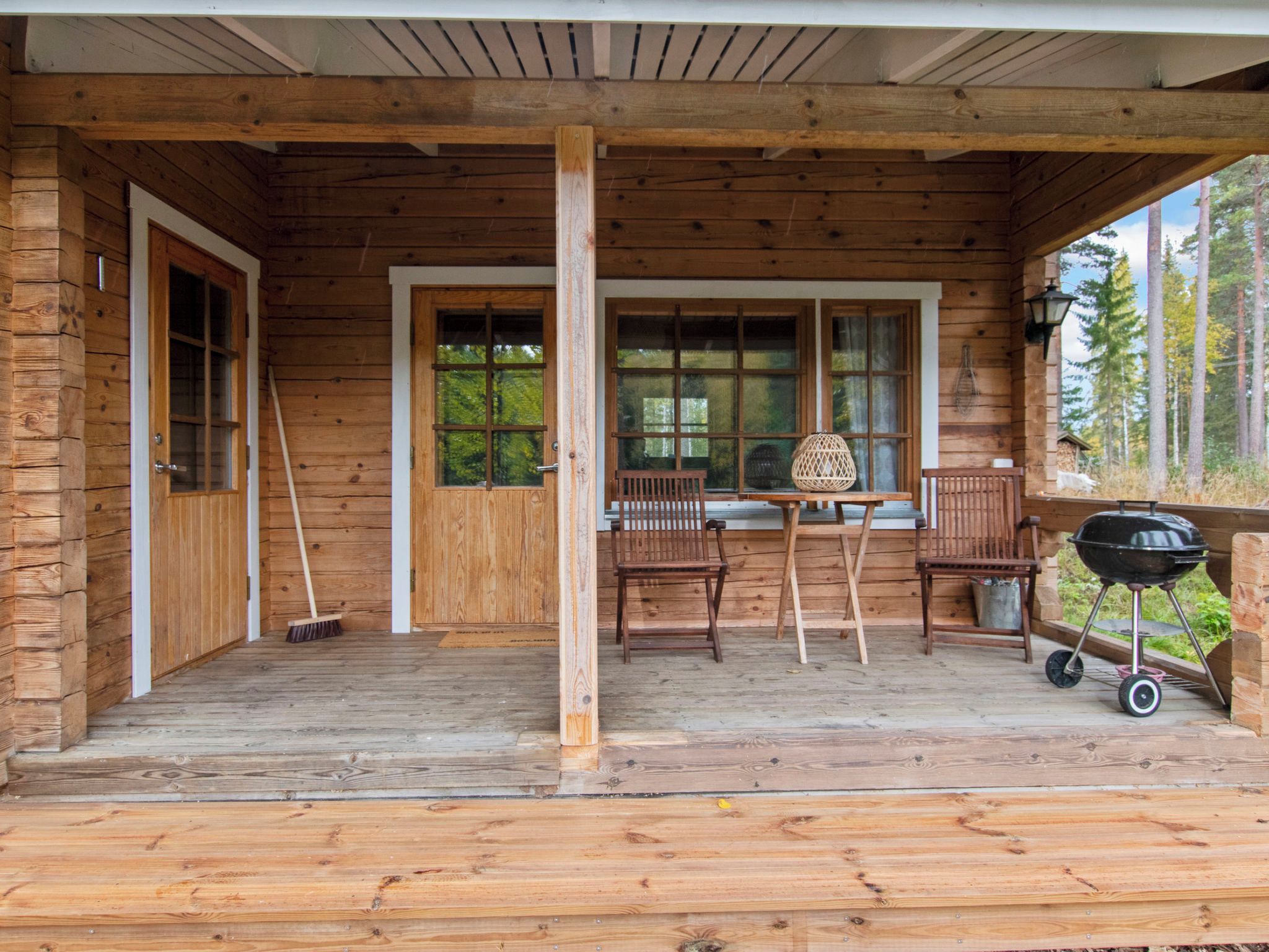 Une cabane en bois avec une véranda, une table à manger, des chaises, un barbecue et une fenêtre donnant sur une forêt.