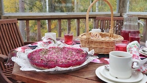 A wooden table with a glass pie on a lace doily, a wicker basket, a pitcher of red drink, and a white cup on a wooden deck with a view of trees. - Koskimökki, vaikon loma 3 by Interhome (Kaavi)