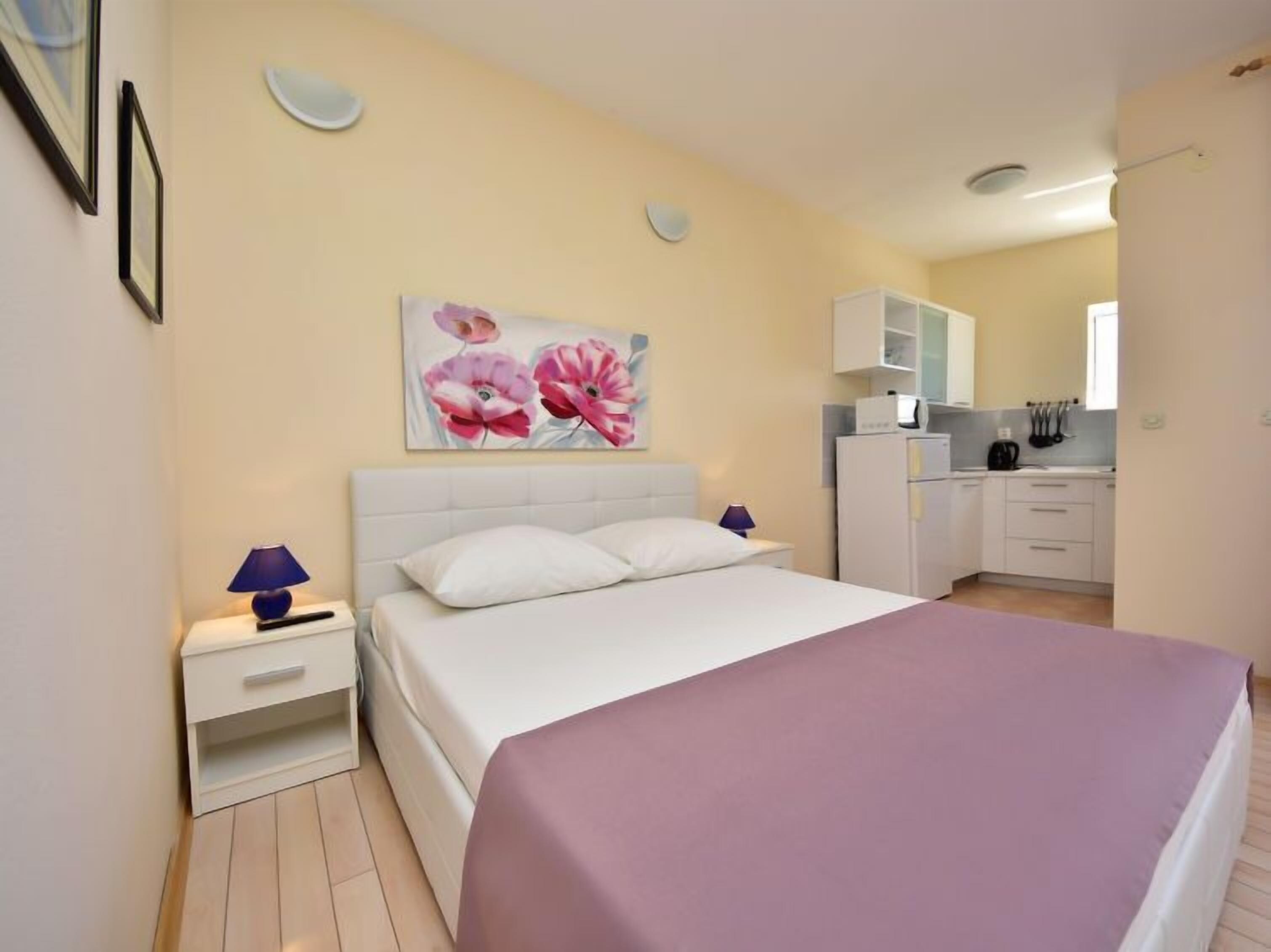 A bedroom with a bed, a nightstand, a lamp, a painting of flowers, and a kitchenette in the background.