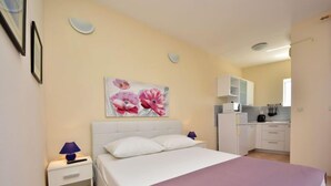 Een slaapkamer met een bed, een nachtkastje, een lamp, een bloemschilderij en een kitchenette op de achtergrond.