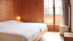 A wooden-paneled bedroom with a large bed, a nightstand, a window with curtains, and a view of the outdoors. - Le Geteillon 4 by Interhome (Villars)