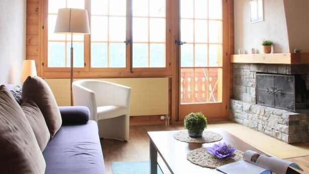A cozy living room with a stone fireplace, a sofa, a chair, and large windows. - Le Geteillon 4 by Interhome (Villars)