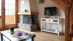 A cozy living room with a stone fireplace, wooden beams, a television on a white cabinet, and a window with a view. - Le Geteillon 4 by Interhome (Villars)