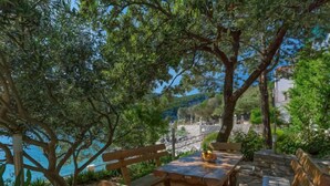 Une table et des bancs en bois sont installés à l’extérieur, offrant une vue sur la mer et les collines en arrière-plan.