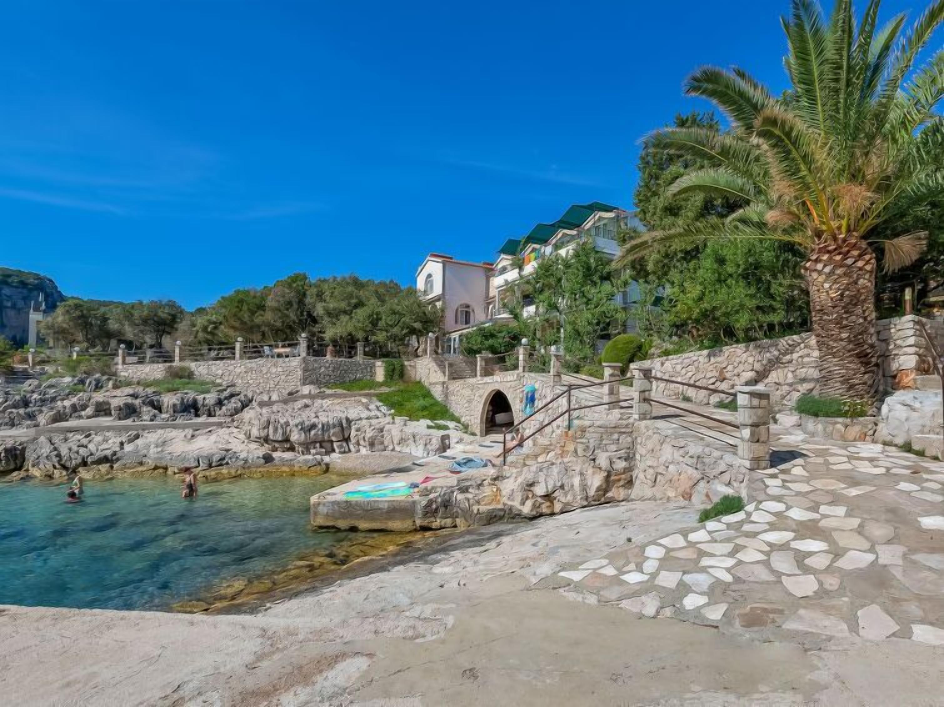 A coastal area with a clear blue sky, rocky terrain, a stone bridge, and a building with a green roof.
