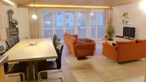 A modern living room with a dining area, featuring a large wooden table, orange sofas, a TV, and a large window with curtains.