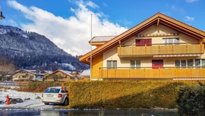 A two-story chalet with a wooden balcony, set against a mountainous backdrop.