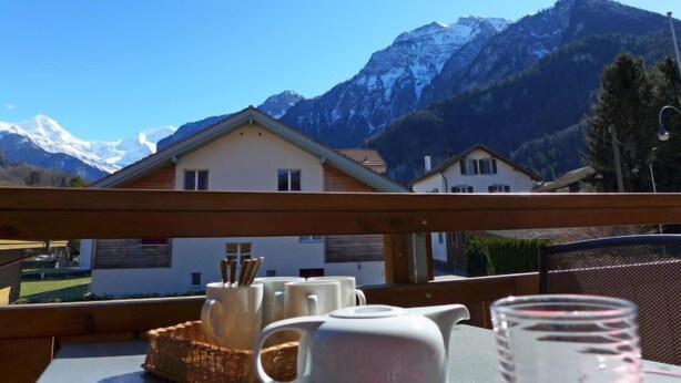 A balcony with a table set for breakfast, overlooking a mountain landscape with houses and snow-capped peaks.