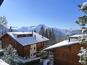 Una cabaña de madera con techo inclinado, rodeada de árboles cubiertos de nieve y montañas al fondo.