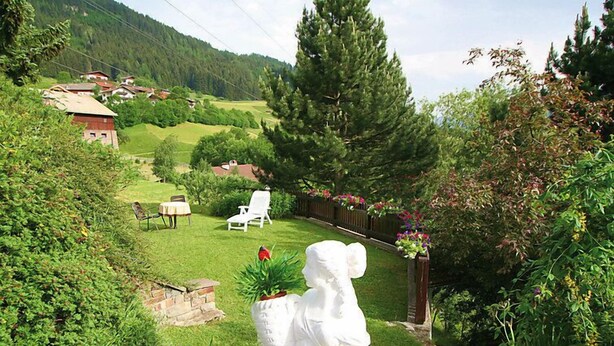 Un jardín con una estatua, césped, cerca de madera y casas al fondo.