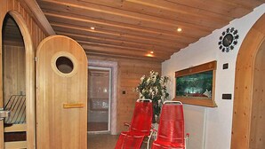 Una sala de sauna con paredes y puerta de madera, dos sillas rojas, una ventana con vistas y un reloj de pared.