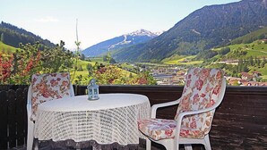 Una mesa redonda con mantel de ganchillo y sillas con motivos florales, con vista a un paisaje montañoso y un pueblo en la falda.