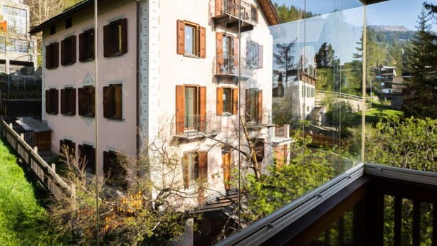 A traditional chalet with a balcony, surrounded by greenery and a clear view of the mountains.
