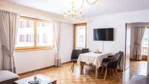 A modern living room with a dining area, wooden parquet flooring, a chandelier, and a flat-screen TV mounted on the wall.