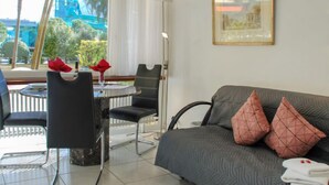 A modern living room with a grey sofa, black chairs, and a small table. There is a window with a view of greenery and a building outside. - Residenza Lido Appartment 008 by Interhome (Locarno)