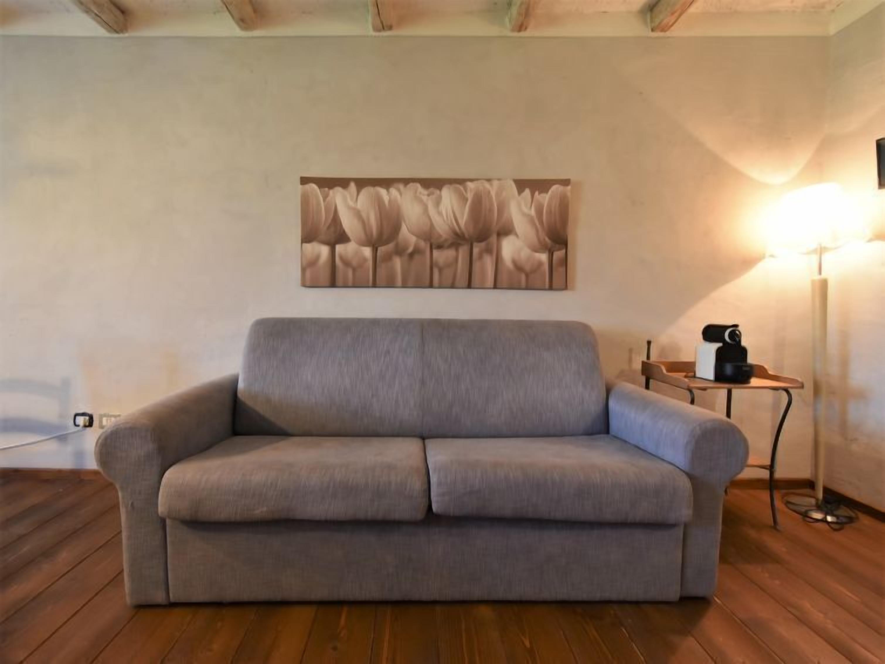 Un sofá gris en una habitación con suelo de madera, un cuadro abstracto colgado en la pared y una lámpara de pie.