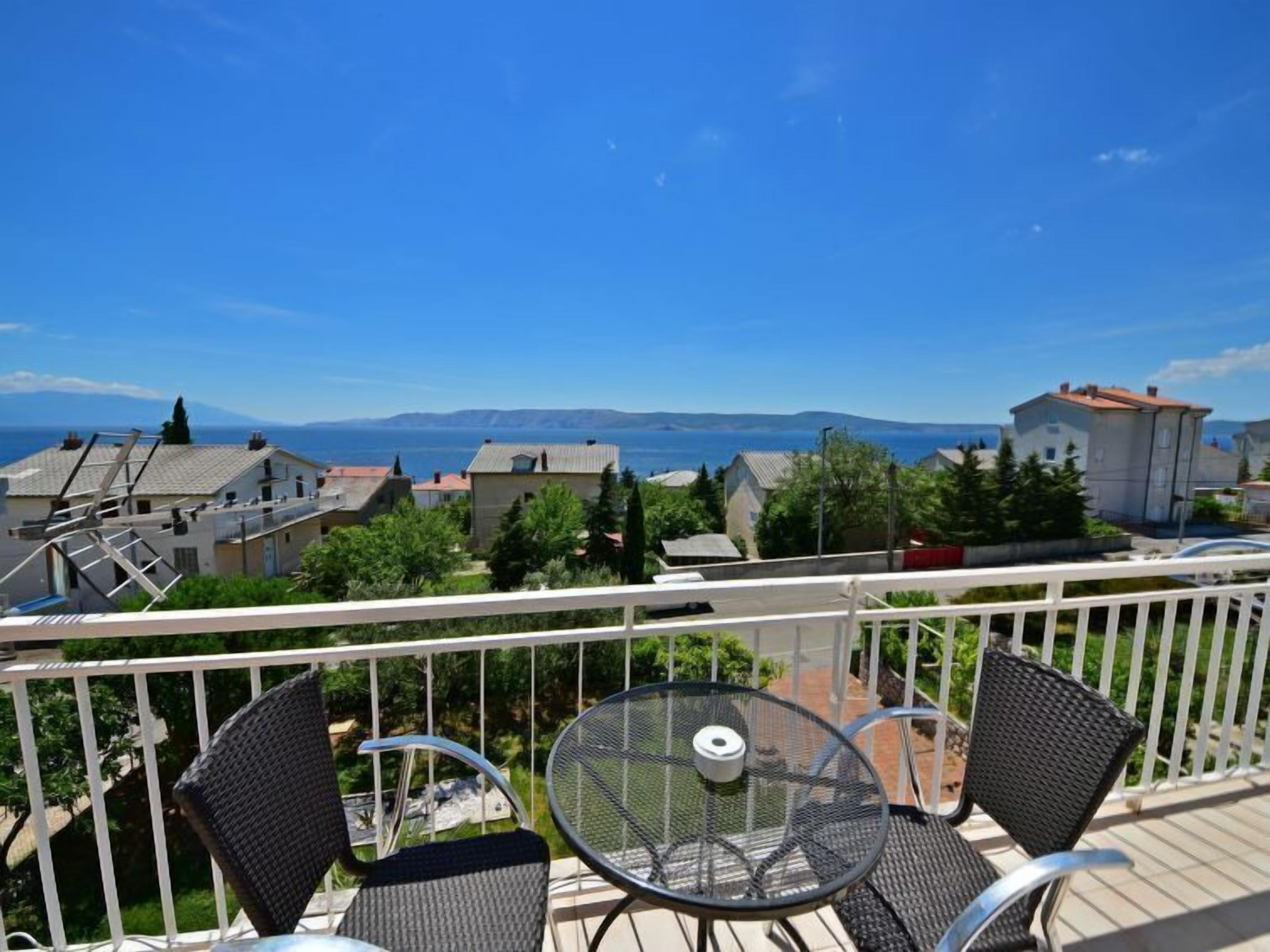 Ein Balkon mit Tisch und Stühlen mit Blick auf eine Küstenstadt, Häuser und einen klaren, blauen Himmel.