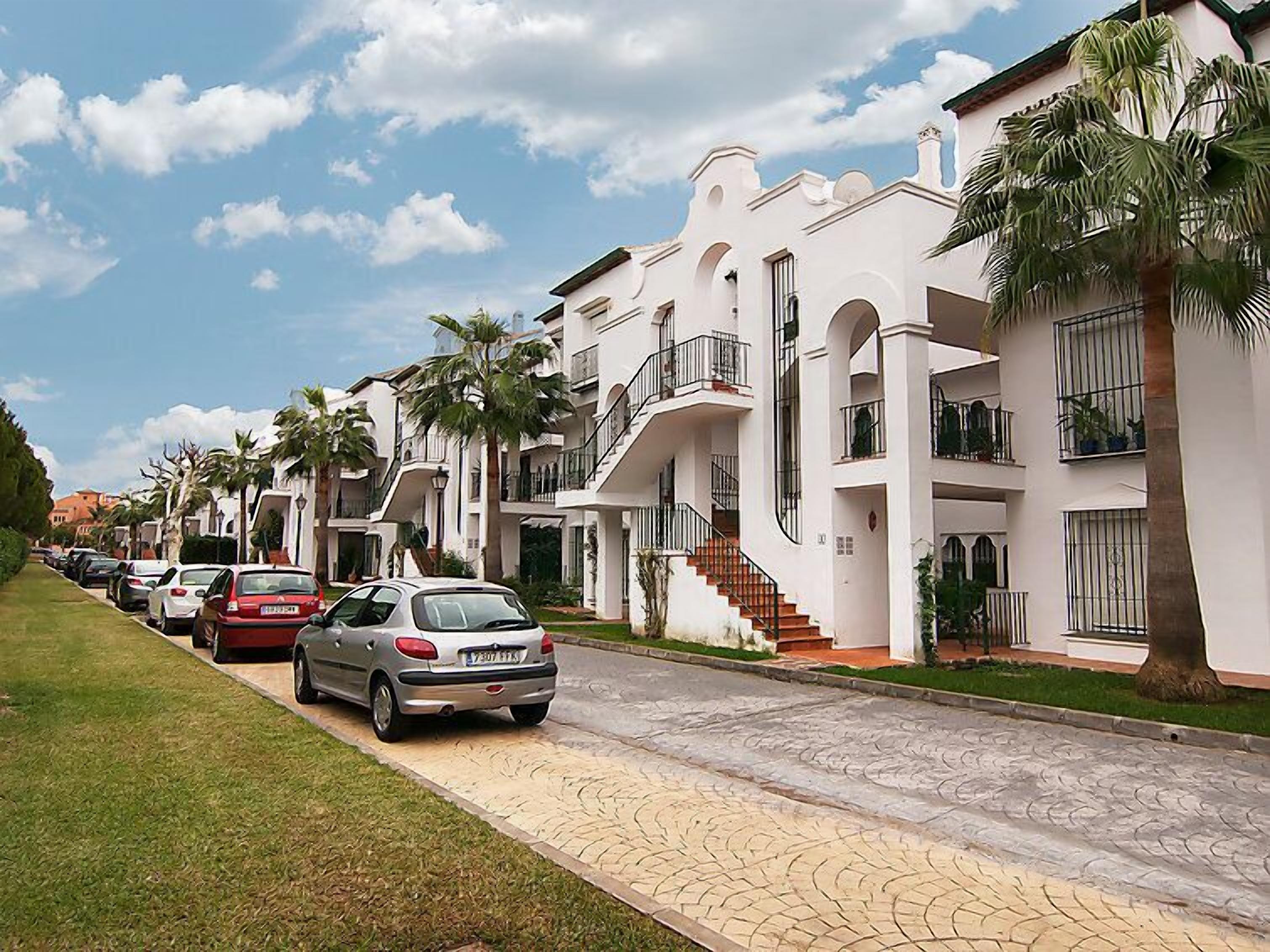 Una hilera de edificios residenciales blancos de varios pisos, con balcones, coches aparcados y palmeras.