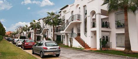 Una hilera de edificios residenciales blancos de varios pisos, con balcones, coches aparcados y palmeras.