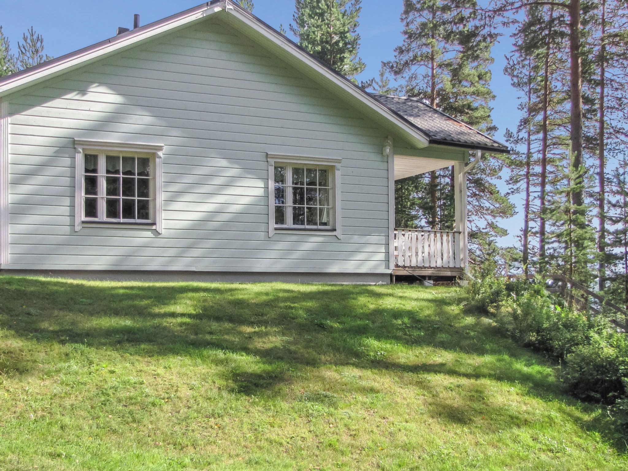 A single-story house with a porch, surrounded by a grassy yard and tall pine trees.