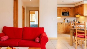 A living room with a red sofa, a dining area with wooden chairs, and a kitchen with wooden cabinets and a microwave. - Les Cimes Blanches 102 A by Interhome (Nendaz)