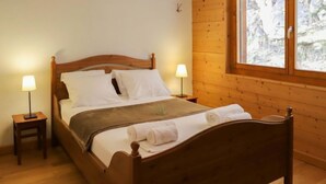 A wooden bed with white linens, two bedside tables with lamps, and a window with a view of trees. - Les Cimes Blanches 102 A by Interhome (Nendaz)