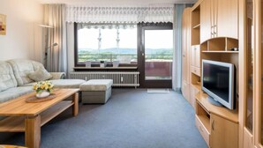 A modern living room with a sofa, a wooden coffee table, a TV on a wooden stand, and a window with a view of hills. - Kiefer (A907) by Interhome (Lahnstein (Koblenz))