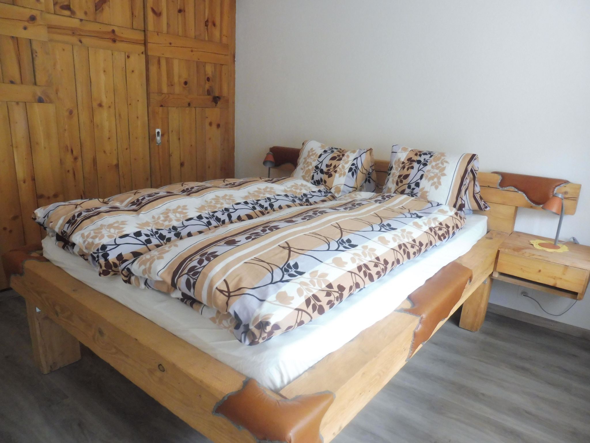 Ein hölzernes Bett mit einem gemusterten Bettbezug und Kissen, ein Nachttisch aus Holz mit einer Lampe sowie ein Holzschrank.