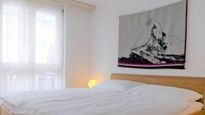 A neatly made bed in a room with a large window and a mountain painting on the wall. - Apartment Roger by Interhome (Zermatt)
