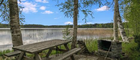 A wooden picnic table with benches, a stone fireplace, and birch trees in a lakeside setting.