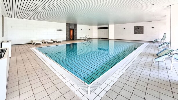 An indoor swimming pool with tiled flooring, a sloped ceiling, and lounge chairs along the sides. - Baumblick (B610) by Interhome (Lahnstein (Koblenz))