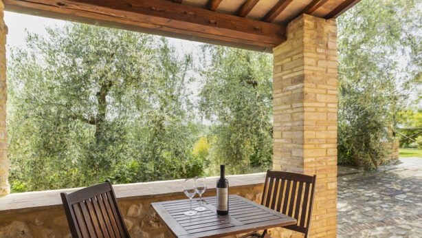 Un espacio de estar al aire libre cubierto, con pared de piedra y sillas de madera, con vistas a un jardín con árboles.